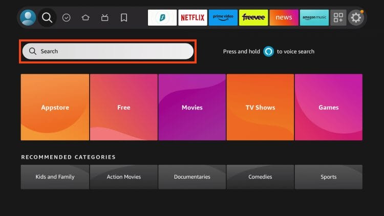 This image shows the home screen of an Amazon Fire TV Stick interface, focusing on the search feature. The search bar is highlighted with an orange border near the top of the screen, indicating where users can type or use voice input to look for apps, movies, TV shows, or games. Above the search bar are shortcut icons and popular streaming app tabs like Netflix, Prime Video, Freevee, News, and Amazon Music.