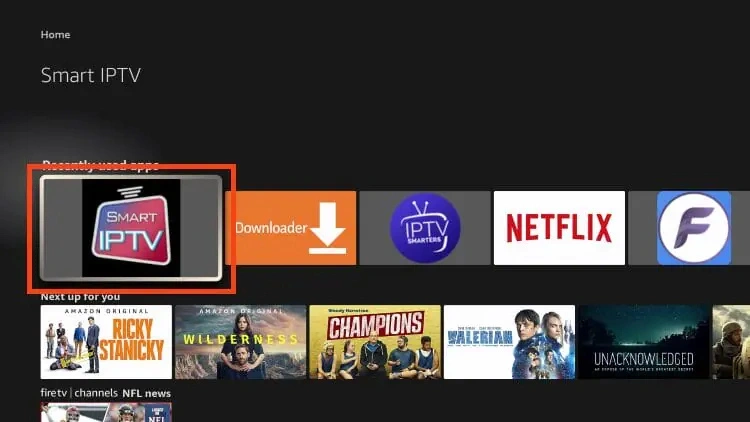 This image shows the FireStick home screen with the Smart IPTV app highlighted under the "Downloaded apps" section. Here are the key elements visible: The Smart IPTV icon is prominently highlighted on the left, showing that it has already been installed and is ready to launch. Other apps installed on the device are visible in a horizontal row, including: Downloader (used to sideload Smart IPTV) IPTV Smarters Netflix Filelinked or FileSynced-type app (logo on the far right) Below the apps row, there’s a “Next up for you” section suggesting Amazon Prime content like Ricky Stanicky, Wilderness, and Champions.