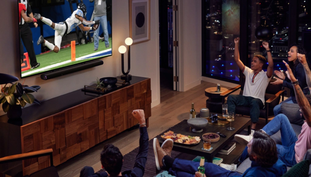 A group of friends cheering while watching an American football game on a large flat-screen TV in a stylish high-rise apartment at night. The room features a wooden media console, snacks on the table, and a city view through the window.