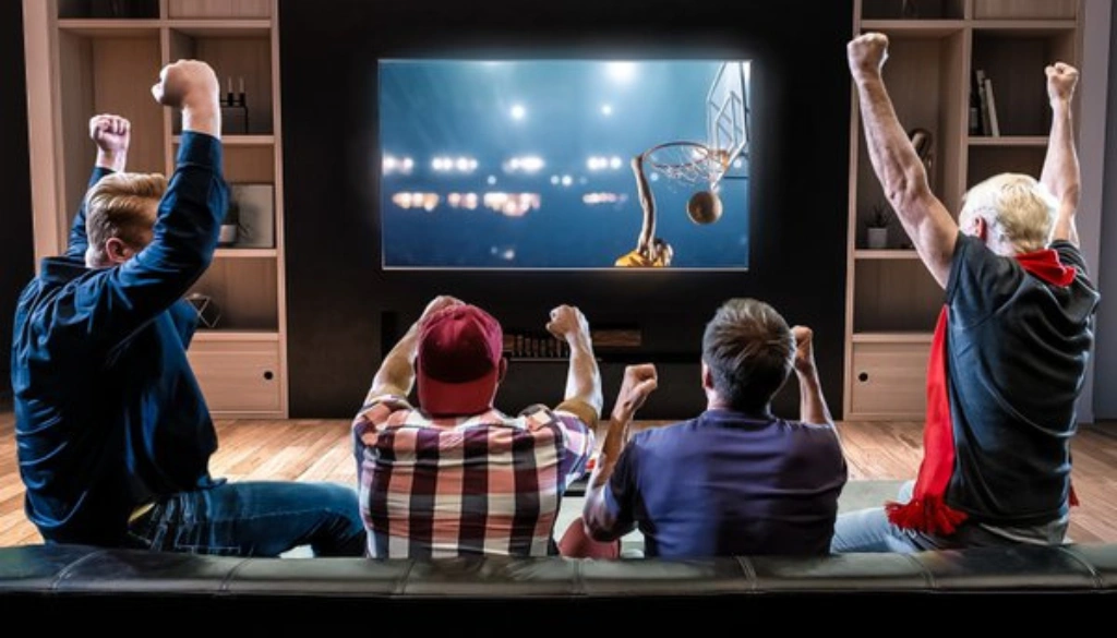 Group of excited friends cheering while watching a basketball game on a large TV screen in a modern living room. The scene captures a thrilling sports moment, with a slam dunk happening on screen and fans raising their arms in celebration.