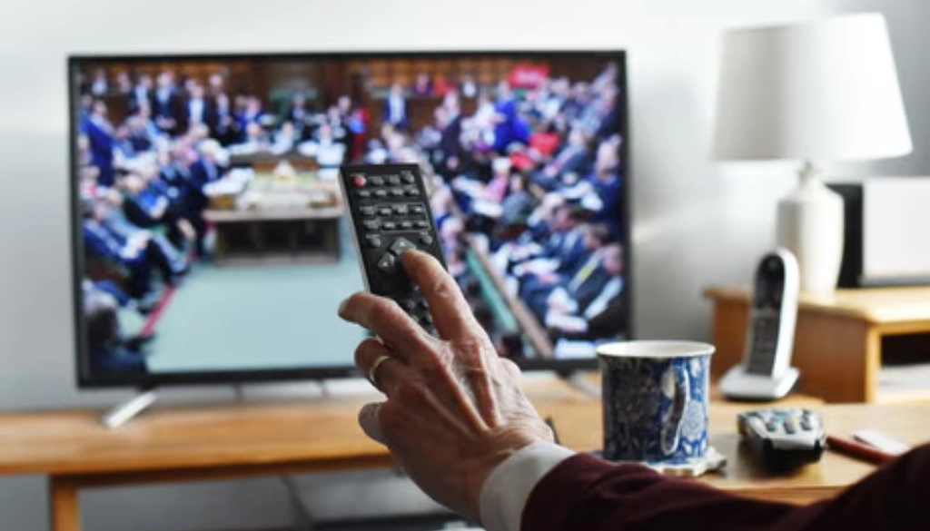 A person holding a TV remote while watching a government or political broadcast on a flat-screen television. The scene is set in a cozy living room with a mug, remote, and lamp on a wooden table.